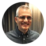 Portrait of a smiling man with glasses and short hair, wearing a dark button-up shirt, standing in front of a neutral background.