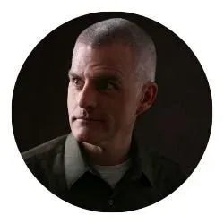 Portrait of a man with short hair, wearing a dark shirt, against a dark background.