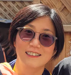 Person wearing sunglasses and smiling, standing outdoors with a wooden fence background.