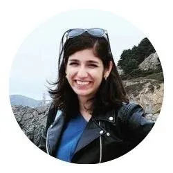 Smiling woman with dark hair wearing sunglasses and a black jacket outdoors in a rocky landscape.