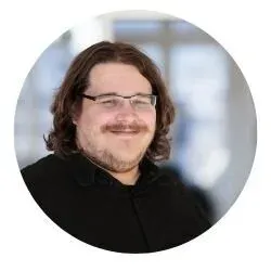 A man with shoulder-length brown hair, glasses, and a beard, smiling in an indoor setting with large windows.