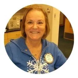A woman smiling in a laboratory or office setting, wearing a blue jacket with winter-themed designs and a name tag.