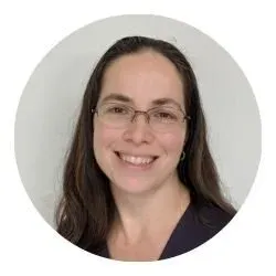 Portrait of a woman with long dark hair, glasses, and a friendly smile, wearing a dark blazer, against a plain light background.