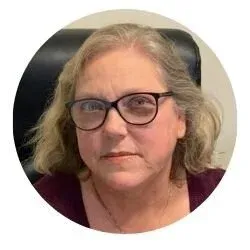 A woman with shoulder-length blonde hair, glasses, and a serious expression