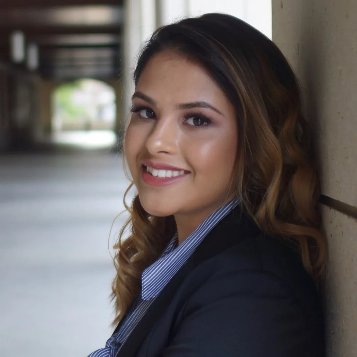 A young woman with long, wavy hair, wearing a business suit, smiling and leaning against a wall in a corridor.