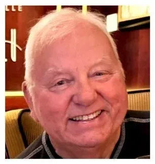 Close-up of a smiling elderly man with light skin and white hair, sitting indoors in a restaurant or cafe.
